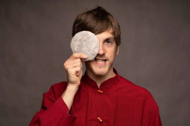 Genç yakışıklı, uzun boylu, zayıf, kahverengi saçlı, bir gözünü pu erh çay kekiyle kaplayan gri arka planda kırmızı gömlekli adam.