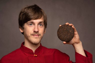 Genç yakışıklı, uzun boylu, zayıf, kahverengi saçlı, kameraya bakan, gri arka planda kırmızı gömlekli, pu erh çayı tutan beyaz adam.