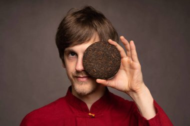 Genç yakışıklı, uzun boylu, zayıf, kahverengi saçlı, sol gözünü kaplayan gri arka planda kırmızı gömlekli, pu erh çay keki olan beyaz adam.