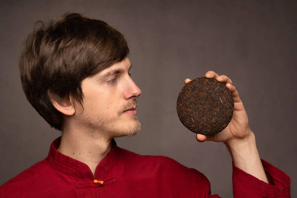 Young handsome tall slim white man with brown hair examining opened pu erh tea cake in red shirt on grey background
