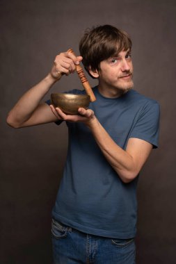 Genç yakışıklı, uzun boylu, zayıf, kahverengi saçlı, gri arka planda mavi gömlekli, kafasının yanında Tibet çanağı taşıyan beyaz bir adam.