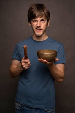 Genç yakışıklı, uzun boylu, zayıf, kahverengi saçlı, gri arka planda Tibet şarkı kasesi taşıyan beyaz bir adam.