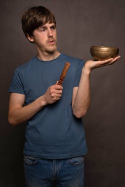 Genç, uzun boylu, zayıf, kahverengi saçlı, düşünceli, gri arka planda mavi tişörtlü Tibet şarkıcısı.