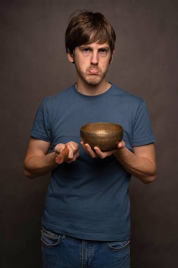 Genç yakışıklı, uzun boylu, zayıf, kahverengi saçlı, gri arka planda Tibet şarkı kasesi tutan, sinirli beyaz adam.