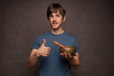 Genç yakışıklı, uzun boylu, zayıf, kahverengi saçlı, gri arka planda Tibet şarkı kasesi tutan, baş parmağını kaldıran beyaz adam.