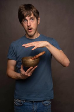 Genç yakışıklı, uzun boylu, zayıf, kahverengi saçlı, gri arka planda mavi tişörtlü, elini Tibet şarkı kasesinin üzerinde gezdiren beyaz adam.