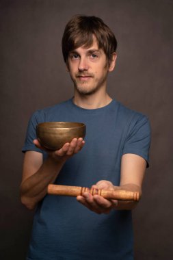 Genç yakışıklı, uzun boylu, zayıf, kahverengi saçlı, gri arka planda Tibet şarkı kasesi taşıyan beyaz bir adam.