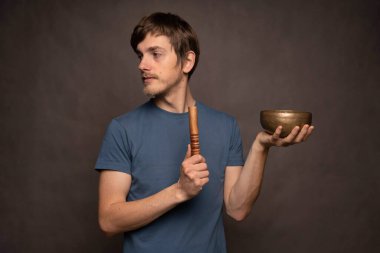 Genç yakışıklı, uzun boylu, zayıf, kahverengi saçlı, gri arka planda mavi gömlekli, Tibet şarkı kasesi tutan beyaz adam.