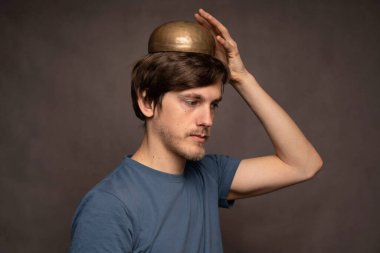 Genç yakışıklı, uzun boylu, zayıf, beyaz, kahverengi saçlı, kafasında mavi gömlekli, gri arka planda Tibet müzik kasesi olan adam.