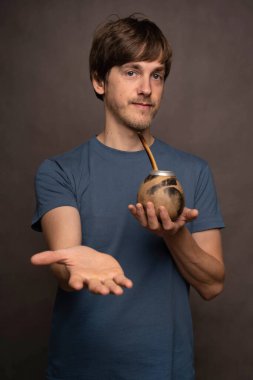 Genç, uzun boylu, zayıf, kahverengi saçlı, mat bardağı tutan, gri arka planda mavi gömlekli genç bir adam.