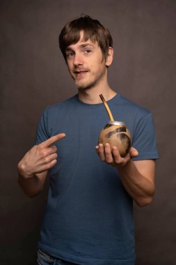 Genç yakışıklı, uzun boylu, zayıf, kahverengi saçlı, gri arka planda mavi gömlekli, mat bardağı gösteren beyaz adam.