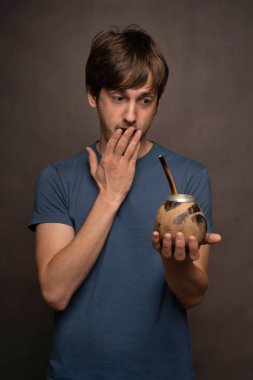 Genç, uzun boylu, zayıf, kahverengi saçlı, mat bardağı tutan gri arka planda, ağzını mavi gömlekle kaplayan beyaz bir adam.