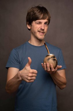 Genç yakışıklı, uzun boylu, zayıf, kahverengi saçlı, baş parmak kaldıran, gri arka planda mavi tişörtlü, jest yapan beyaz adam.