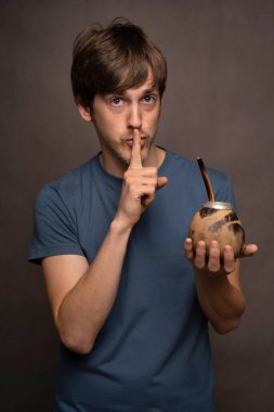 Genç yakışıklı, uzun boylu, zayıf, kahverengi saçlı, mat bardağı tutan, dudakları dudaklarında mavi gömlekli gri arka planda genç bir adam.