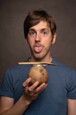 Genç yakışıklı, uzun boylu, zayıf, kahverengi saçlı, mat bardağı tutan, gri arka planda mavi gömlekli, beyaz bir adam.