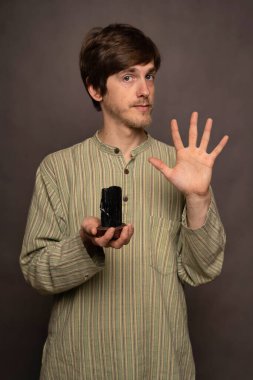 Genç yakışıklı, uzun boylu, zayıf, kahverengi saçlı, elini havada tutan gri arka planda siyah kristal gömlekli adam.