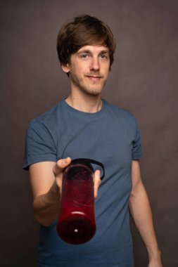 Genç yakışıklı, uzun boylu, zayıf, kahverengi saçlı beyaz adam gri arka planda gri gömlekli, güvenli bir şekilde smoothie ikram ediyor.