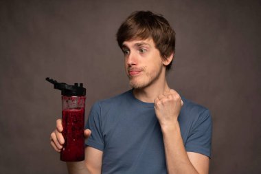 Genç yakışıklı, uzun boylu, zayıf, kahverengi saçlı, gri arka planda gri gömlekli, smoothie şişesi için heyecanlanan beyaz adam.