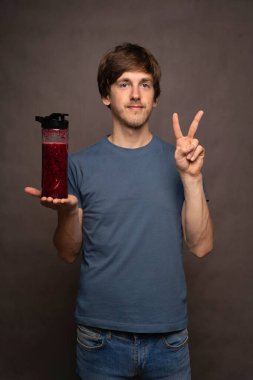 Genç yakışıklı, uzun boylu, zayıf, kahverengi saçlı, smoothie şişeli beyaz adam gri arka planda gri gömlekli bir barış işareti yapıyor.