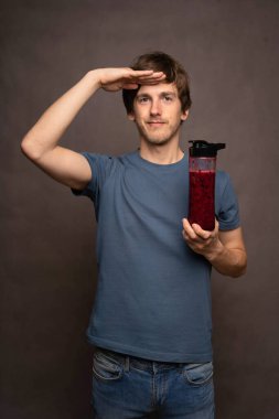 Gri arka planda gri gömlekli smoothie şişesini tutarken kahverengi saçlı, uzun boylu, zayıf beyaz bir adam selam veriyor.
