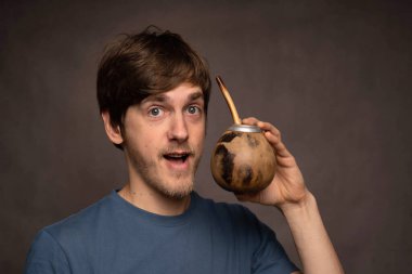 Genç yakışıklı, uzun boylu, zayıf, kahverengi saçlı, gri arka planda mat fincanıyla poz veren beyaz adam.