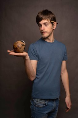 Genç yakışıklı, uzun boylu, zayıf, kahverengi saçlı, mat bardağını sağ elinde tutan gri arka planda gri gömlekli adam.