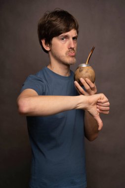 Genç yakışıklı, uzun boylu, zayıf, kahverengi saçlı, mat bardağı tutan, gri arka planda gri tişörtlü, beyaz bir adam.