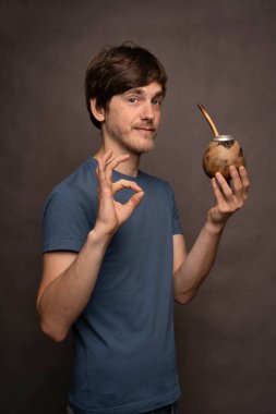 Genç yakışıklı, uzun boylu, zayıf, kahverengi saçlı, elinde mat fincanı olan beyaz adam gri arka planda gri gömleğiyle 