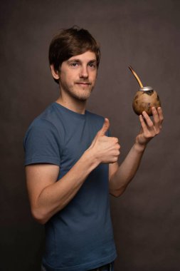 Genç, uzun boylu, zayıf, kahverengi saçlı, mat bardağı tutan, gri arka planda gri gömlekli, beyaz bir adam.