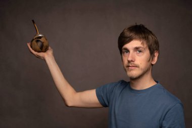 Genç yakışıklı, uzun boylu, zayıf, kahverengi saçlı, mat bardağı tutan gri gömlekli, gri arka planlı beyaz adam.