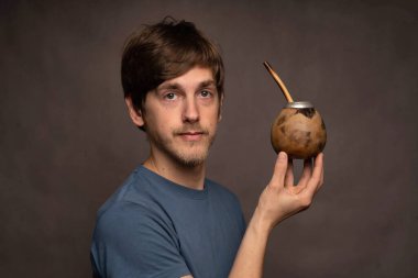 Genç yakışıklı, uzun boylu, zayıf, kahverengi saçlı, mat bardağı tutan gri gömlekli, gri arka planlı beyaz adam.