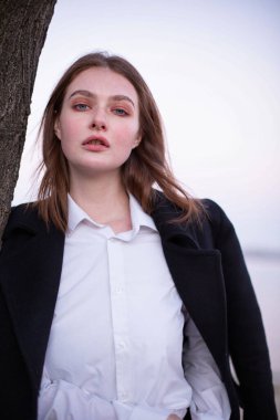 Uzun düz saçlı, koyu renk ceketli, arkasında göl olan beyaz Avrupalı bir kadının portresi.