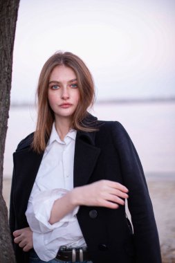 Uzun düz saçlı, koyu renk ceketli, arkasında göl olan beyaz Avrupalı bir kadının portresi.