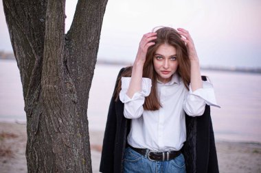 Kumsaldaki ağacın yanında siyah ceketli, uzun düz saçlı, beyaz Avrupalı kadın.