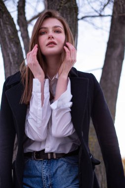 Uzun düz saçlı, beyaz Avrupalı kadın. Yüzüne yakın elleri olan, kumsaldaki ağacın yanındaki koyu renk paltolu kadın.