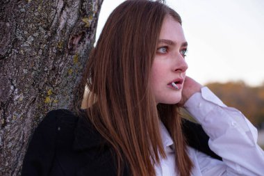 Uzun düz saçlı, beyaz Avrupalı kadın, ağacın yanında koyu renk ceketli.