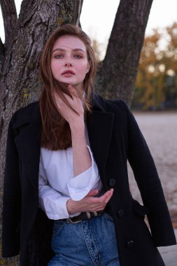 Uzun düz saçlı, beyaz Avrupalı kadın, ağacın yanında koyu renk ceketli.