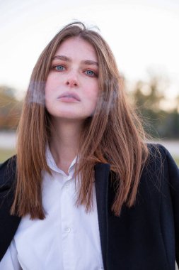 Önünde sigara dumanı olan, sonbaharda kumsalda koyu renk ceketli, uzun düz saçlı beyaz Avrupalı kadının portresi.