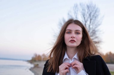 Uzun düz saçlı, beyaz Avrupalı bir kadının portresi. Sonbaharda kumsalda yakasını koyu bir ceketle düzeltiyor.