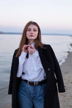 Uzun düz saçlı, beyaz Avrupalı kadın, sonbaharda kumsalda koyu renk ceketiyle yakasını düzeltiyor.