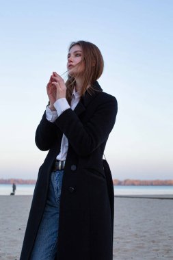 Uzun düz saçlı, beyaz Avrupalı kadın. Sonbaharda kumsalda elleri katlanmış, koyu renk ceketli.