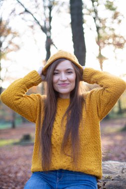 Güneşli sonbahar ormanlarında sarı kazaklı Avrupalı beyaz kadın başının arkasında el ele tutuşuyor.