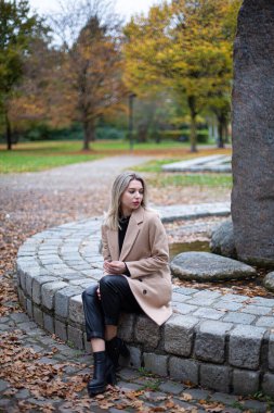 Parkta sarı saçlı, bej rengi sonbahar ceketli, taş çitin üzerinde oturan, ciddi, beyaz Avrupalı genç bir kadın.