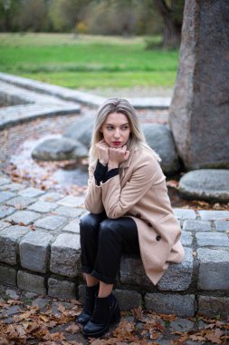 Parkta sarı saçlı Avrupalı beyaz bir kadın. Taş çitin üzerinde oturuyor. Elleri çenesinin altında bej rengi sonbahar ceketi giyiyor.