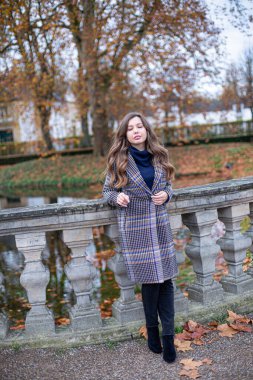 Sonbahar parkında taş çitin yanında duran uzun kıvırcık saçlı trençkotlu genç kadın.