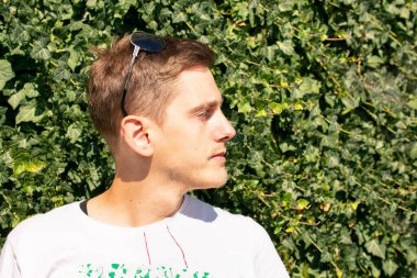 Kafasında güneş gözlüğü olan, yeşil çitin önüne bakan, beyaz gömlekli, kahverengi saçlı, yakışıklı beyaz bir adam.