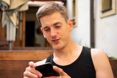 Siyah fanilalı kahverengi saçlı yakışıklı beyaz adam terasta telefonuna gülümsüyor.