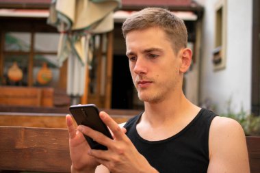 Siyah fanilalı kahverengi saçlı yakışıklı beyaz adam terasta telefonuna bakıyor.