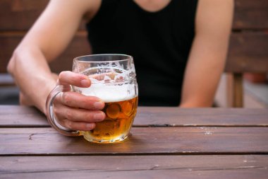 Siyah fanilalı genç beyaz adam bar terasında bira tutuyor.