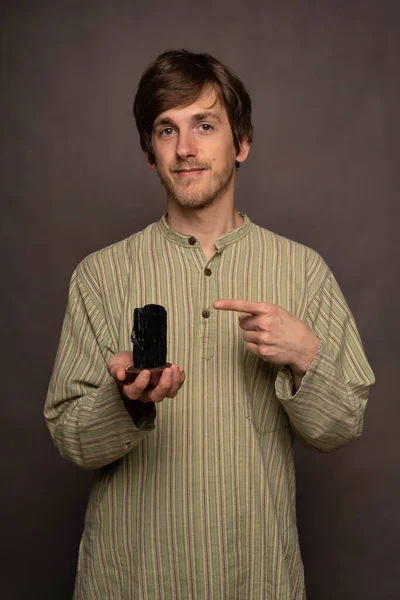 Genç, uzun boylu, zayıf, kahverengi saçlı, gri arkaplanda siyah kristali gösteren beyaz bir adam.